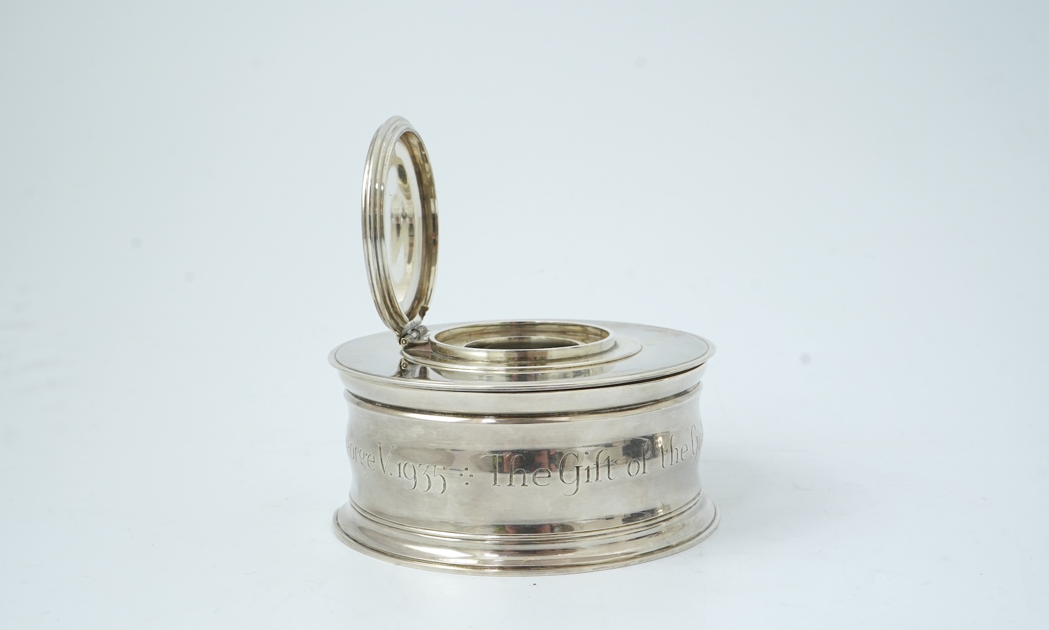 A George V silver circular inkwell, by Sebastian Garrard, commemorating the Silver Jubilee of King George V, the die stamped cover designed by George Kruger Gray featuring the Goldsmiths Company Coat of Arms
